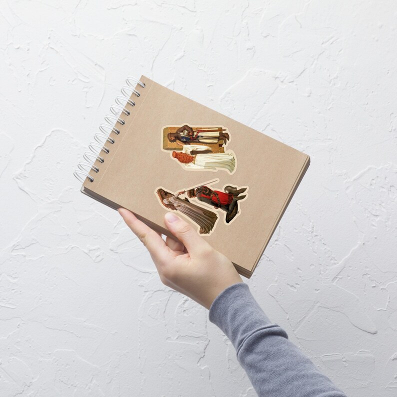 May include: A spiral-bound notebook with three stickers featuring vintage illustrations of figures in historical attire. The stickers depict a knight in armor, a woman in a white dress, and a figure in red and black clothing.