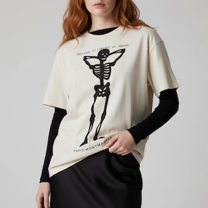 Puede incluir: Camiseta color crema con un dise&ntilde;o de esqueleto negro. La camiseta tiene el texto "SOUVENIR DU CABARET DU NEANT" y "PARIS-MONTMARTRE". La camiseta de manga corta se lleva sobre una camiseta negra de manga larga.