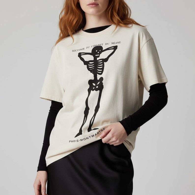 Puede incluir: Camiseta color crema con un dise&ntilde;o de esqueleto negro. La camiseta tiene el texto "SOUVENIR DU CABARET DU NEANT" y "PARIS-MONTMARTRE". La camiseta de manga corta se lleva sobre una camiseta negra de manga larga.