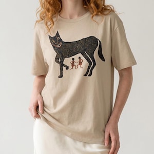 May include: Beige t-shirt featuring a black illustration of a smiling cat-like creature with two small figures beneath it. The graphic has a textured appearance. The shirt has short sleeves and a crew neck.