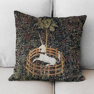 May include: A throw pillow with a tapestry design featuring a white unicorn in a circular fence, surrounded by a lush green forest with colorful flowers.
