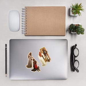 May include: Three stickers featuring medieval art scenes are displayed on a laptop. One shows a kneeling knight, another depicts a woman in a long dress, and the third shows a figure in a coffin. A mouse, pen, and notebook are also visible.