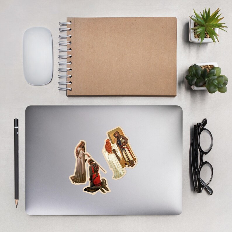 May include: Three stickers featuring medieval art scenes are displayed on a laptop. One shows a kneeling knight, another depicts a woman in a long dress, and the third shows a figure in a coffin. A mouse, pen, and notebook are also visible.