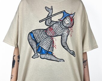 Pode incluir: Camiseta bege com um gráfico de um cavaleiro medieval em cota de malha, com detalhes azuis e vermelhos. O cavaleiro é retratado em uma pose dinâmica, segurando uma arma. A camiseta é uma peça de roupa casual.