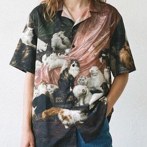 May include: A short-sleeve button-down shirt featuring a detailed, multi-colored print of numerous cats in a classical setting with draped pink fabric. The shirt has a notched collar, a straight hem, and a dark, painterly background aesthetic.