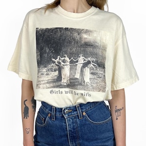 May include: Cream-coloured t-shirt with a black and white image of figures in a circle, and the text "Girls will be girls" printed below. The shirt is worn with blue denim jeans.