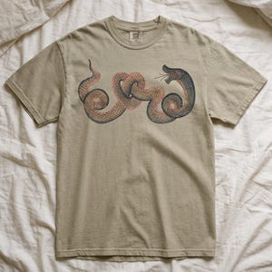 May include: A short-sleeved crew neck t-shirt in a muted beige colour. The front features a graphic illustration of a coiled snake with dark grey scales, a red underbelly, and an open mouth showing fangs. The snake is positioned horizontally.