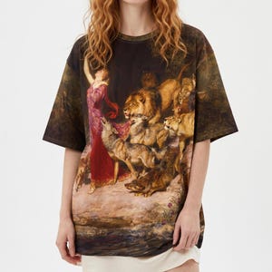 May include: A short-sleeve t-shirt featuring a full-color graphic print of a classical painting. The artwork depicts a figure in a long red dress standing among a group of lions and wolves in a dark, forest-like setting with earthy tones.
