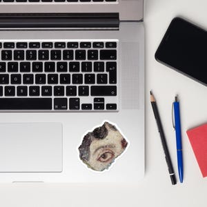 May include: A sticker featuring a watercolor painting of an eye. The eye is brown with a white and black background. The sticker is on a white surface next to a laptop, a phone, a pencil, a pen, and a red notepad.