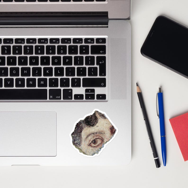 May include: A sticker featuring a watercolor painting of an eye. The eye is brown with a white and black background. The sticker is on a white surface next to a laptop, a phone, a pencil, a pen, and a red notepad.