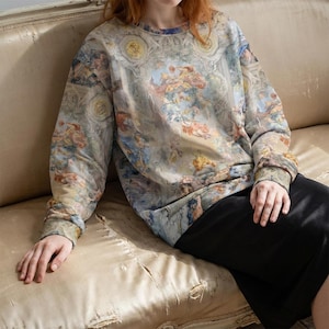 May include: A crew neck sweatshirt with a Renaissance-style painting print in shades of blue, beige, and peach. The long-sleeved top features a detailed design reminiscent of classical art, offering a unique and artistic aesthetic.