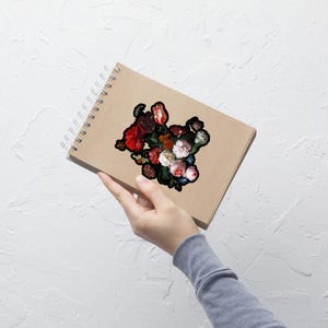 Pu&ograve; includere: Un taccuino marrone a spirale con un adesivo floreale raffigurante un bouquet di fiori rossi, bianchi e rosa. L'adesivo ha un bordo nero. Il taccuino &egrave; tenuto da una persona.