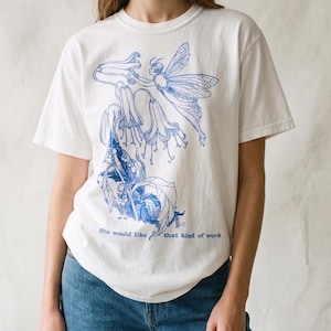 Peut inclure: Un t-shirt blanc à manches courtes avec une impression graphique bleue représentant une fée ailée interagissant avec des fleurs en forme de cloche. Le texte "She would like that kind of work" est imprimé en bleu sous l'illustration.