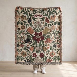 May include: A rectangular woven throw blanket with a floral pattern in shades of red, green, beige, and brown. The blanket has a fringed edge and is held up by a person. The floral design is symmetrical.