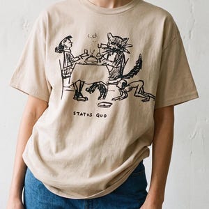 Pode incluir: Uma t-shirt bege de manga curta com um gráfico de linhas pretas que mostra uma pessoa e um gato grande sentados a uma mesa com uma pequena criatura por baixo. O texto "STATUS QUO" está impresso a preto sob a ilustração frontal.