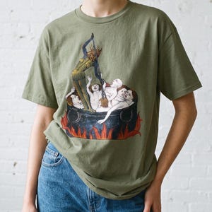 以下が含まれることがあります： オリーブグリーンの半袖Tシャツ。大きな黒い大釜の中に複数の人物が入っているグラフィックプリントが施されています。角のある黄色い肌の生き物が鍋の中に立ち、中の人々の上に長い金属製の道具をかざしています。