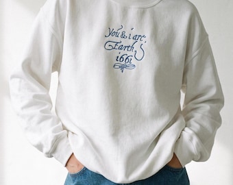Op de afbeelding: Een wit sweatshirt met ronde hals en donkerblauw borduursel op de borst met de tekst "You & i are, Earth, 1661" en een decoratieve krul eronder. Het kledingstuk heeft lange mouwen en een geribbelde kraag, boorden en zoom.
