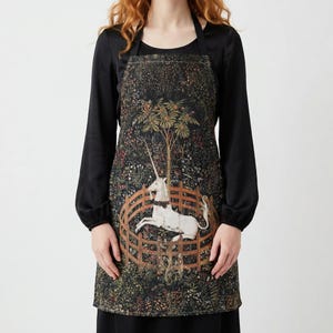 May include: A black apron featuring a tapestry design with a white unicorn inside a wooden fence, set against a backdrop of a lush forest with a tree. The apron has a halter neck and a rectangular shape.