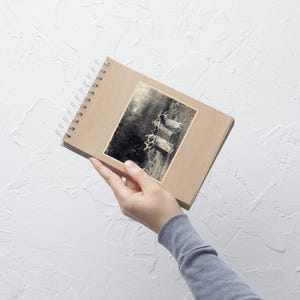 May include: A spiral-bound notebook with a kraft paper cover. A black and white sticker of a vintage image of people on swings is affixed to the cover. The notebook is held by a person wearing a gray long-sleeved shirt.