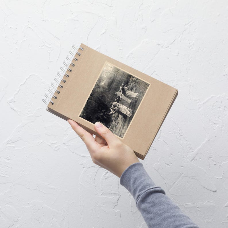 May include: A spiral-bound notebook with a kraft paper cover. A black and white sticker of a vintage image of people on swings is affixed to the cover. The notebook is held by a person wearing a gray long-sleeved shirt.