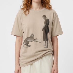 May include: A beige short-sleeved t-shirt featuring a monochromatic graphic print of a person standing in simple clothing and a dog lying on the ground. The illustration is rendered in shades of gray and brown, centered on the front of the garment.