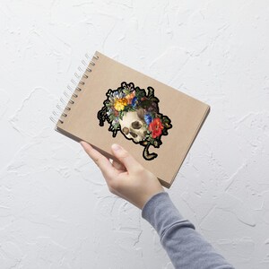 May include: A black and white illustration of a skull with a floral crown. The skull is surrounded by colorful flowers, including red, yellow, blue, and green.