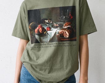 Pode incluir: Uma t-shirt verde-azeitona com uma impressão retangular da pintura Still Life of the Remnants of a Meal with a Lunging Cat de Alexandre-François Desportes, retratando um gato a tentar alcançar comida numa mesa com fruta e carne.