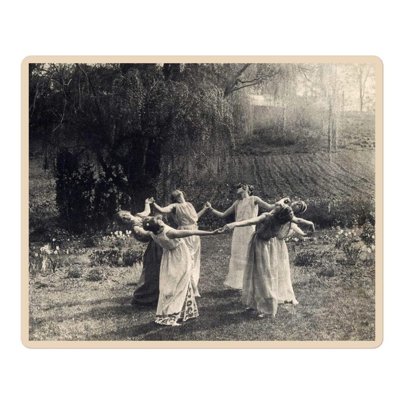 May include: A vintage black and white photograph depicts a group of people in long dresses holding hands in a circle. They are in a grassy area with trees and flowers in the background. The image has a sepia tone.