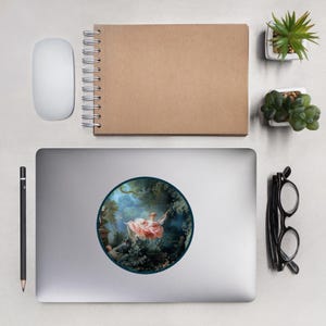 May include: A round sticker featuring a classical painting of a woman on a swing in a garden. The artwork is framed by a dark blue border and set against a silver laptop. Other items include a white mouse, a brown notebook, and small potted plants.
