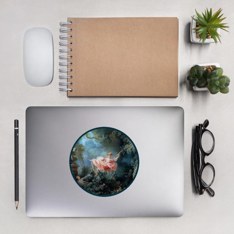 May include: A round sticker featuring a classical painting of a woman on a swing in a garden. The artwork is framed by a dark blue border and set against a silver laptop. Other items include a white mouse, a brown notebook, and small potted plants.