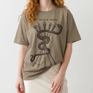 Op de afbeelding: Olijfgroen t-shirt met een zwarte illustratie van een slang en pijlen, met de tekst "VIS NESCIA VINCI". Het shirt heeft korte mouwen en een losse pasvorm. Het ontwerp is op de voorkant gedrukt.