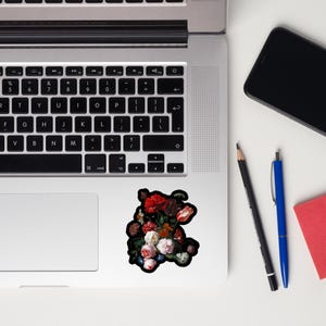 Pu&ograve; includere: Un adesivo floreale con un bouquet di fiori rossi, rosa e bianchi con foglie verdi. L'adesivo &egrave; su un laptop, accanto a una matita nera, una penna blu e un blocco note rosso. I fiori hanno uno sfondo scuro.