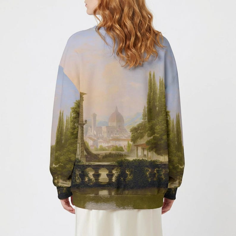 May include: A long-sleeved sweatshirt with a scenic landscape painting. The artwork shows a European cityscape with a domed building, tall trees, and water. The sweatshirt has a light blue and green colour palette.