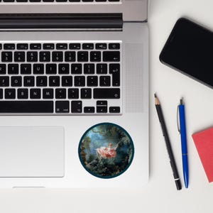 May include: A round sticker featuring a classical painting of a woman in a pink dress on a swing. The artwork is set in a lush green forest with a blue sky background. The sticker is on a laptop.