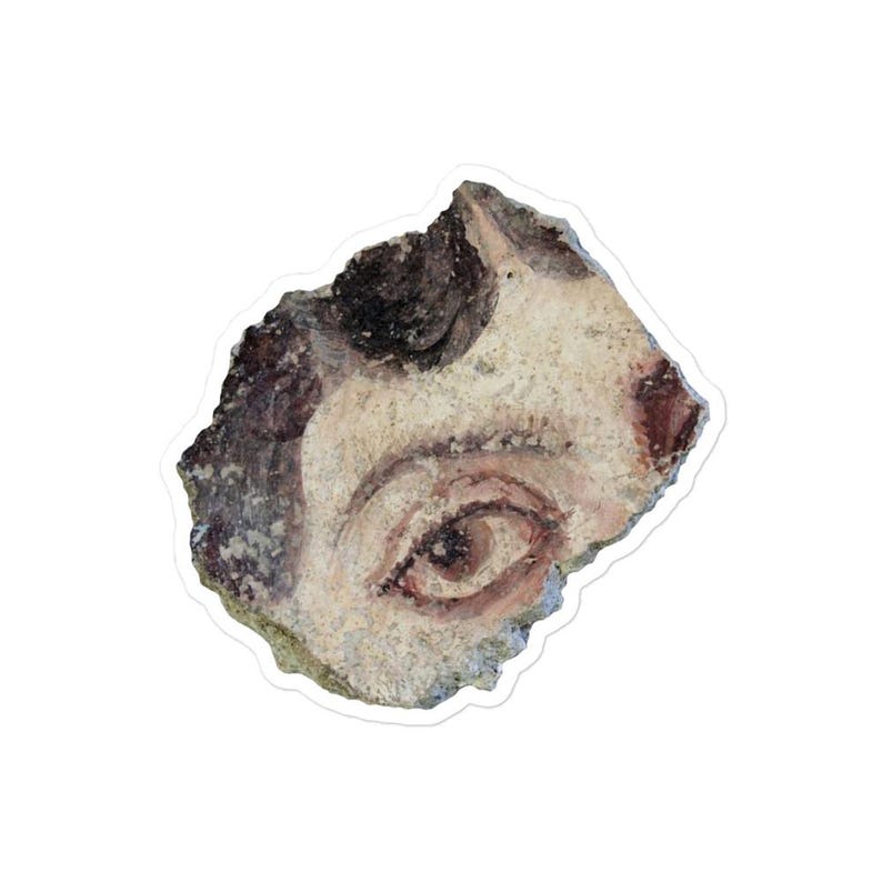 May include: A sticker featuring a fragment of ancient artwork. The artwork depicts a human eye and part of a face, painted in shades of brown, red, and beige. The sticker has a white border.