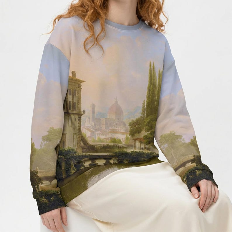 May include: A light blue sweatshirt featuring a scenic landscape painting of a European city with a river, buildings, and trees. The long-sleeved top has black cuffs and a light blue crew neck.