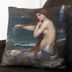 Puede incluir: Una pintura de una sirena con el pelo largo y rojo, sentada en una costa rocosa. Est&aacute; sujet&aacute;ndose el pelo con una mano y mirando hacia un lado. El agua es azul y hay rocas y algas en el fondo.