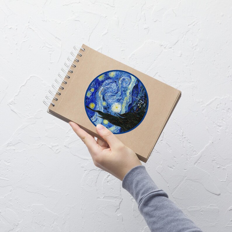 May include: A spiral-bound notebook with a brown cover. The cover features a circular print of Van Gogh's "Starry Night" painting, with swirling blue and yellow brushstrokes. The notebook is held against a white background.