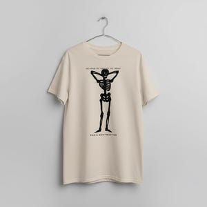Puede incluir: Camiseta beige con un dise&ntilde;o de esqueleto negro con los brazos detr&aacute;s de la cabeza. El texto "SOUVENIR DU CABARET DU NEANT" est&aacute; encima del esqueleto, y "PARIS-MONTMARTRE" debajo.