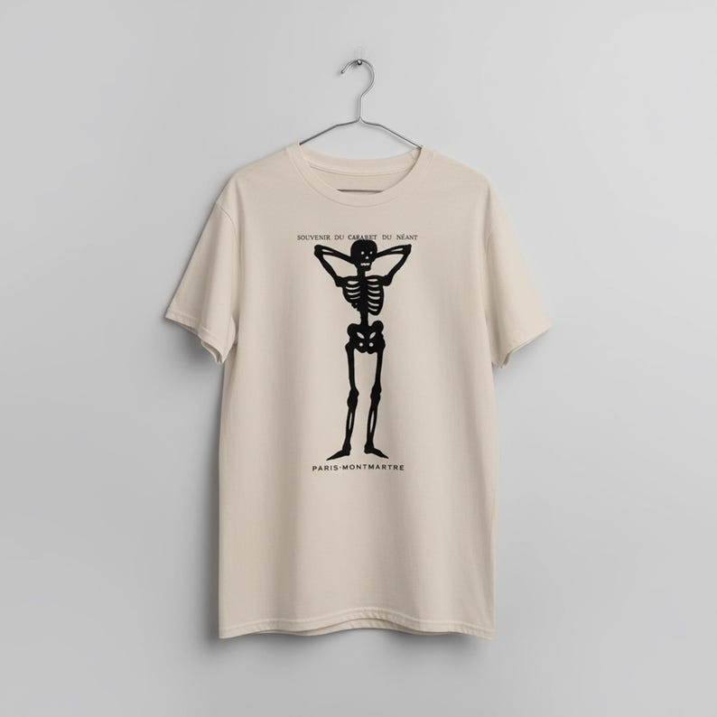 Puede incluir: Camiseta beige con un dise&ntilde;o de esqueleto negro con los brazos detr&aacute;s de la cabeza. El texto "SOUVENIR DU CABARET DU NEANT" est&aacute; encima del esqueleto, y "PARIS-MONTMARTRE" debajo.