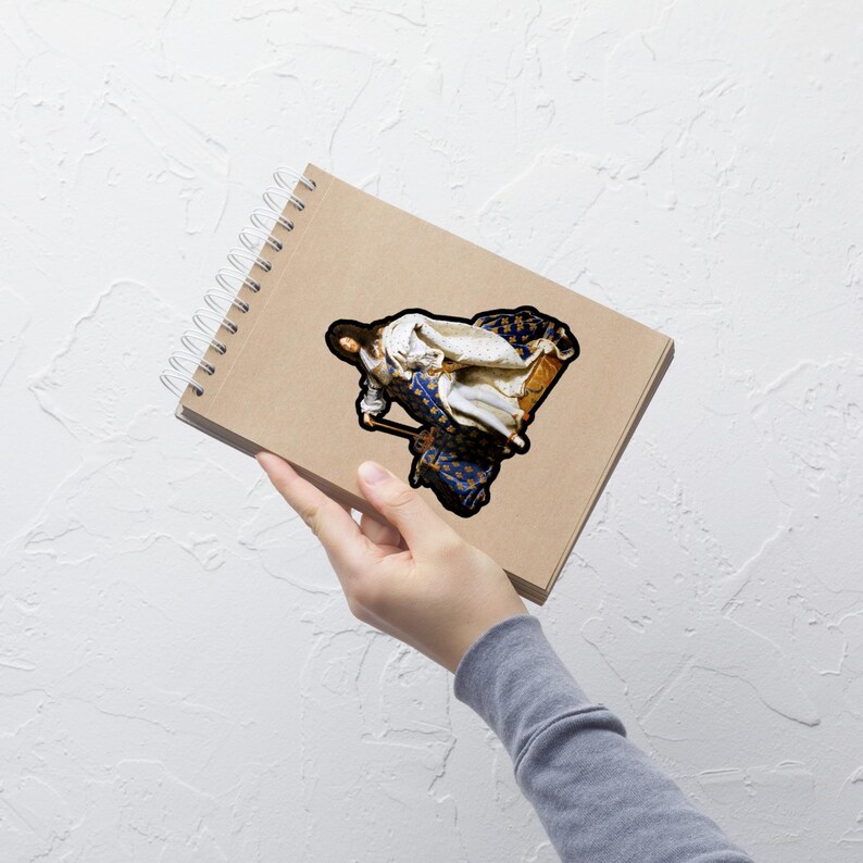 以下が含まれることがあります： 椅子に寄りかかっている人物の古典絵画をあしらったステッカー。このアートワークは、白と金のローブを着た人物を、青と金の模様の背景に描いています。ステッカーは、スパイラル綴じのノートに貼られています。