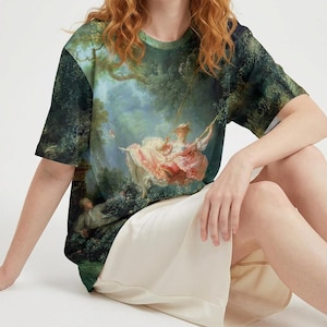 May include: A short-sleeved t-shirt displaying a classical painting of a woman on a swing in a garden setting. The artwork uses green, pink, and white tones, with detailed trees and figures. The shirt is worn by a person sitting on a white surface.