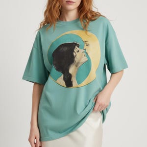 Op de afbeelding: Een zeegroen t-shirt met een vintage illustratie van een vrouw die naar een halve maan kijkt. De maan heeft een gezicht en kust de vrouw. De afbeelding staat in een cirkelvormig kader.