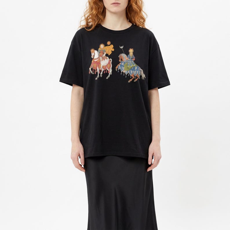 Puede incluir: Camiseta negra con un gr&aacute;fico colorido de dos figuras a caballo, con coronas y s&iacute;mbolos, sobre un fondo de sol y luna. La camiseta tiene un corte holgado y de gran tama&ntilde;o.