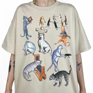 Puede incluir: Camiseta beige con una ilustración caprichosa de estilo medieval de varios gatos en diferentes poses. Los gatos están representados en tonos de azul, gris, naranja y negro, algunos con coronas o tocando instrumentos.