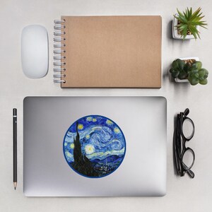 May include: A laptop with a sticker featuring Van Gogh's "Starry Night" painting. The sticker is a circular design with a blue border, showcasing the swirling night sky, stars, and a dark silhouette of a village and cypress tree. Other items include a mouse, notebook, and glasses.