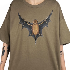 May include: Olive green t-shirt with a bat illustration. The bat has a brown body, black wings, and a detailed face. The shirt has a crew neck and short sleeves. The t-shirt is a relaxed fit.