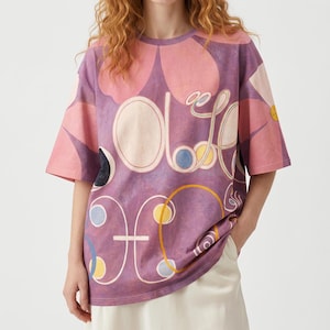 Puede incluir: Una camiseta de manga corta en tonos morados y rosas con un gran estampado floral y gr&aacute;ficos abstractos de l&iacute;neas blancas. El dise&ntilde;o incluye elementos circulares en amarillo, azul y negro, con un texto superpuesto en letra cursiva blanca.