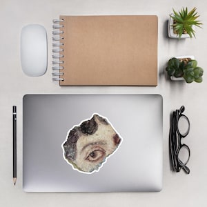 May include: A sticker featuring a fragment of a classical painting, showcasing a detailed eye with brown iris and surrounding facial details. The sticker is irregularly shaped and placed on a laptop, surrounded by office supplies.