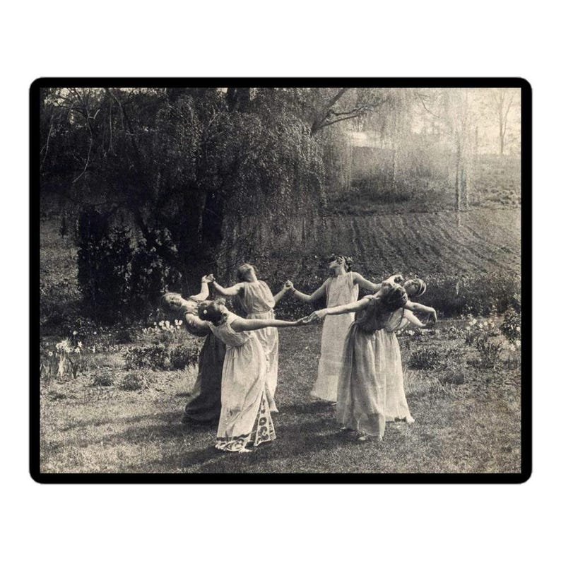 May include: Black and white photograph of a group of people in long dresses dancing in a circle outdoors. The image is set in a grassy area with trees and a field in the background. The people are holding hands.